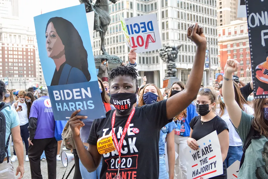 A woman at a rally holds up a Biden Harris sign with her fist raised and a photo of Kamala Harris in the background.